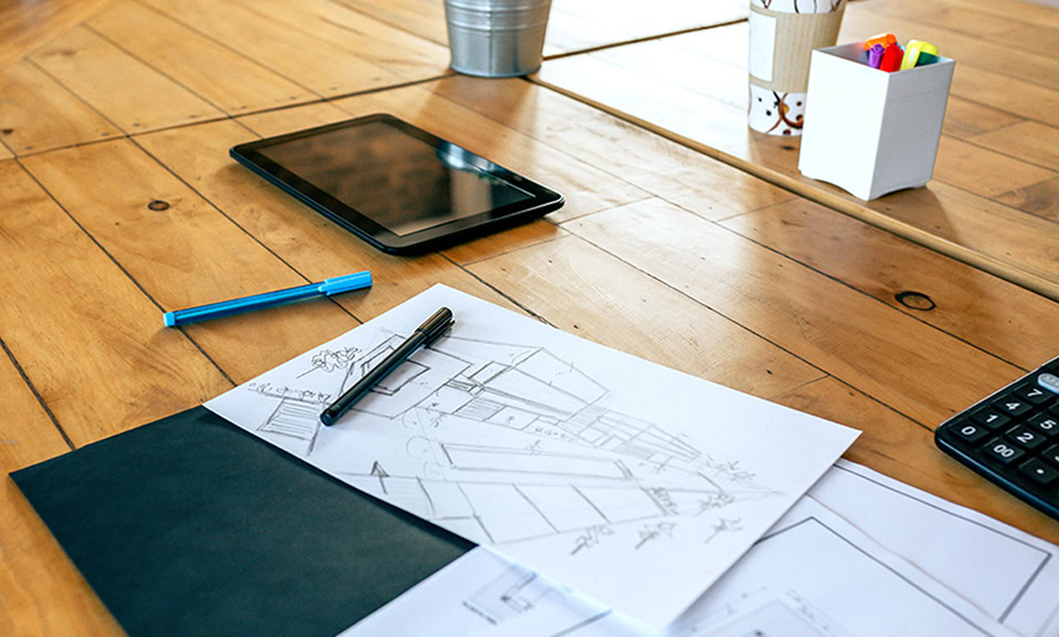 Architectural sketches, a tablet, a pen, a blue pencil, a calculator, and a container with markers are arranged on a wooden table.