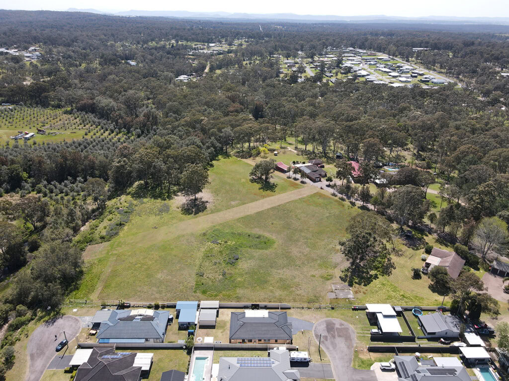 Aerial view of a rural residential area with houses at the bottom, open grassy land in the center, and dense forested areas in the background.