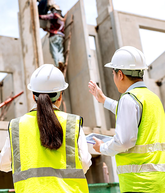 Two construction workers in safety vests and helmets observe and discuss the progress of a building site; one holds a tablet while another worker is seen climbing in the background.
